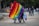 A vibrant scene in a park with a group of people holding large, colorful rainbow flags. In the background, other people are visible walking and chatting. Trees provide a lush backdrop, and there are vendors with carts selling refreshments. The atmosphere is lively and inclusive.