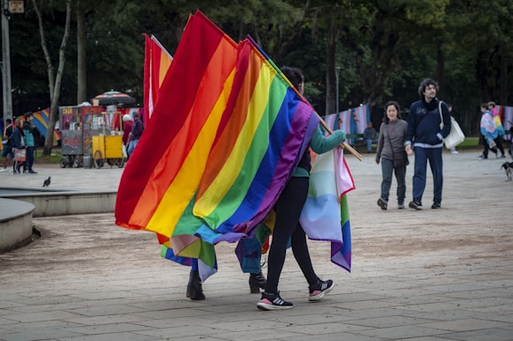 A vibrant scene in a park with a group of people holding large, colorful rainbow flags. In the background, other people are visible walking and chatting. Trees provide a lush backdrop, and there are vendors with carts selling refreshments. The atmosphere is lively and inclusive.