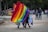 A vibrant scene in a park with a group of people holding large, colorful rainbow flags. In the background, other people are visible walking and chatting. Trees provide a lush backdrop, and there are vendors with carts selling refreshments. The atmosphere is lively and inclusive.