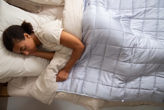 A person is peacefully sleeping in bed, lying on their side. The bed has a light beige pillow and sheets, and a gray quilted blanket covers the sleeper. Their head rests on the pillow, and the soft bedding creates a cozy atmosphere.