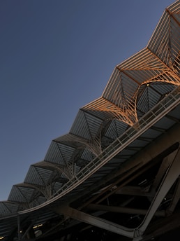An architectural structure with a modern, angular design featuring a series of glass and metal canopies arranged in a repeating pattern. The structure is highlighted by soft, warm light from the setting or rising sun, casting shadows that enhance the geometric lines.