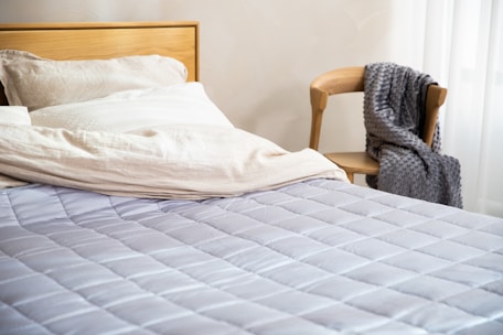 A neatly made bed with a wooden headboard and beige pillows. The bed is covered with a light gray quilted blanket. To the right, there is a wooden chair with a textured gray blanket draped over it. Soft natural light filters through a nearby window with sheer white curtains.