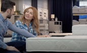 A couple is seated on different mattresses in a store, engaging in conversation. The woman, with long red hair, wears a blue plaid shirt, while the man has short dark hair and a blue shirt. Behind them are shelves displaying rolled-up mattresses and bedding items under soft, dim lighting.