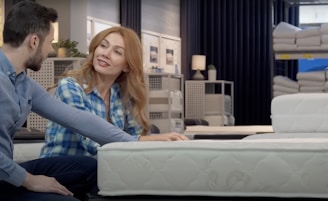 A couple is seated on different mattresses in a store, engaging in conversation. The woman, with long red hair, wears a blue plaid shirt, while the man has short dark hair and a blue shirt. Behind them are shelves displaying rolled-up mattresses and bedding items under soft, dim lighting.