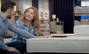 A couple is seated on different mattresses in a store, engaging in conversation. The woman, with long red hair, wears a blue plaid shirt, while the man has short dark hair and a blue shirt. Behind them are shelves displaying rolled-up mattresses and bedding items under soft, dim lighting.