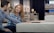 A couple is seated on different mattresses in a store, engaging in conversation. The woman, with long red hair, wears a blue plaid shirt, while the man has short dark hair and a blue shirt. Behind them are shelves displaying rolled-up mattresses and bedding items under soft, dim lighting.