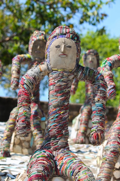 Sculptures made from colorful bangles are displayed outdoors, resembling human figures with abstract faces. The figures are positioned in dynamic poses, surrounded by a lush green backdrop of trees.