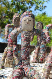 Sculptures made from colorful bangles are displayed outdoors, resembling human figures with abstract faces. The figures are positioned in dynamic poses, surrounded by a lush green backdrop of trees.