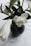 A dark vase holds elegant white flowers, including a large rose and lilies, surrounded by lush green leaves. The arrangement is set against a pale, marble-like background, highlighting the contrast between the soft petals and the sleek vase.