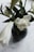 A dark vase holds elegant white flowers, including a large rose and lilies, surrounded by lush green leaves. The arrangement is set against a pale, marble-like background, highlighting the contrast between the soft petals and the sleek vase.