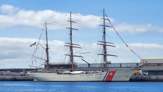 A large sailing ship is docked at a harbor under a partly cloudy sky. The ship has three tall masts, each equipped with rigging and an array of flags. The hull is painted white with a 'U.S. Coast Guard' label, and there's a red, white, and blue stripe near the bow. An American flag is waving at the stern.