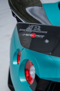 The image features the rear section of a teal sports car, highlighting a carbon fiber spoiler with logos from APR Performance and J-Spec Perf Edition. The glossy teal surface contrasts with the dark carbon fiber material. The design emphasizes the car's aerodynamic and performance-driven features.