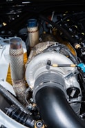 A close-up view of a turbocharger system in an engine bay, featuring metallic tubes and pipes wrapped in heat-resistant material, with the BorgWarner logo visible. The components have a mix of polished and textured surfaces.