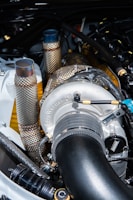 A close-up view of a turbocharger system in an engine bay, featuring metallic tubes and pipes wrapped in heat-resistant material, with the BorgWarner logo visible. The components have a mix of polished and textured surfaces.