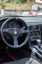 The image depicts the interior of a car, focusing on the steering wheel and dashboard. The steering wheel is a sporty Momo design with a black finish and racing details. The dashboard features multiple gauges, a speedometer, and a tachometer. The center console displays classic car controls, including an aftermarket stereo system. The gear shift is visible, along with some decorative elements such as a sticker near the gear lever.