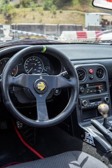 The image depicts the interior of a car, focusing on the steering wheel and dashboard. The steering wheel is a sporty Momo design with a black finish and racing details. The dashboard features multiple gauges, a speedometer, and a tachometer. The center console displays classic car controls, including an aftermarket stereo system. The gear shift is visible, along with some decorative elements such as a sticker near the gear lever.