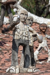 Several sculptures made from mosaic tiles and concrete stand on a surface covered with tiles. The figures are human-like with simplified facial features and are composed of broken ceramic pieces that form their bodies and clothing. The sculptures vary in position, with one prominently holding a cup-like object.