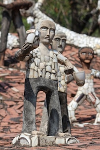 Several sculptures made from mosaic tiles and concrete stand on a surface covered with tiles. The figures are human-like with simplified facial features and are composed of broken ceramic pieces that form their bodies and clothing. The sculptures vary in position, with one prominently holding a cup-like object.