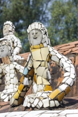 Mosaic sculptures composed of colorful ceramic tiles depict human-like figures. They are placed on a brick surface, and the background features blurred greenery.
