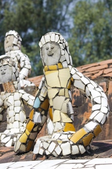 Mosaic sculptures composed of colorful ceramic tiles depict human-like figures. They are placed on a brick surface, and the background features blurred greenery.