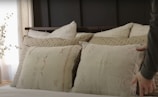 An arrangement of decorative pillows on a bed features varied textures and neutral colors. The bed appears cozy with layered cushions featuring embroidered patterns. A hand is seen adjusting one of the pillows. The background includes soft lighting and a hint of a plant, contributing to a serene bedroom atmosphere.