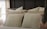 An arrangement of decorative pillows on a bed features varied textures and neutral colors. The bed appears cozy with layered cushions featuring embroidered patterns. A hand is seen adjusting one of the pillows. The background includes soft lighting and a hint of a plant, contributing to a serene bedroom atmosphere.