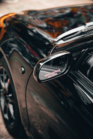 A close-up view of a black car focusing on its side mirror, which contains the humorous text 'OBJECTS IN MIRROR ARE LOSING' in red letters. The car's surface reflects the surroundings, showcasing its glossy finish.