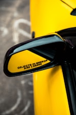 A close-up of a car's side mirror with a humorous message stating 'OBJECTS IN MIRROR ARE LOSING.' The mirror is framed by a bright yellow car body, offering a sleek and sporty appearance.