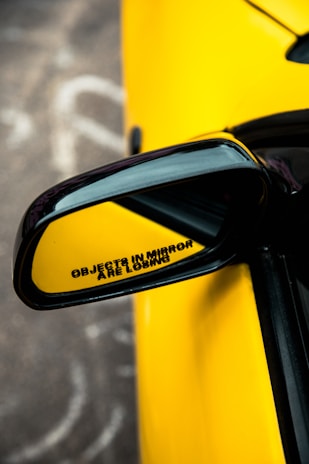 A close-up of a car's side mirror with a humorous message stating 'OBJECTS IN MIRROR ARE LOSING.' The mirror is framed by a bright yellow car body, offering a sleek and sporty appearance.