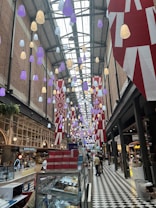 A high-ceilinged indoor market with decorative, multi-colored hanging lights in purple, white, and yellow. The ceiling is made of glass and supported by metal beams. The market features various shops and food stalls, with some people shopping or walking along the black and white checkered floor. Large red and white banners hang from the ceiling.