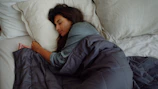 A person lies asleep on a bed, wrapped in a dark grey comforter. Their head rests on a light-colored pillow, and they appear peaceful and relaxed.