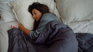 A person lies asleep on a bed, wrapped in a dark grey comforter. Their head rests on a light-colored pillow, and they appear peaceful and relaxed.