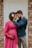 A couple stands close to each other, with the man gently holding the woman's face while she places her hand on her pregnant belly. The woman is wearing a bright pink dress adorned with a white and red flower in her curly hair. The man is dressed in a blue shirt and beige pants. They appear to be standing in front of a white door with brick walls on either side.