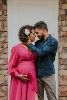 A couple stands close to each other, with the man gently holding the woman's face while she places her hand on her pregnant belly. The woman is wearing a bright pink dress adorned with a white and red flower in her curly hair. The man is dressed in a blue shirt and beige pants. They appear to be standing in front of a white door with brick walls on either side.