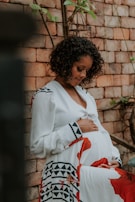 A pregnant woman gently holding her belly while leaning against a brick wall. She is wearing a long white dress with geometric and floral patterns in black and red. Her expression is serene and introspective. Vines and foliage surround the wall, adding a natural element to the scene.