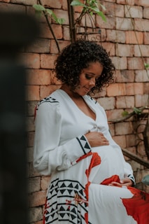 A pregnant woman gently holding her belly while leaning against a brick wall. She is wearing a long white dress with geometric and floral patterns in black and red. Her expression is serene and introspective. Vines and foliage surround the wall, adding a natural element to the scene.