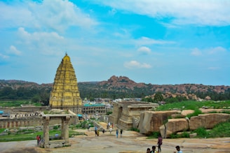 A historical temple with an ornate, towering structure stands prominently against a clear blue sky. Surrounding the temple are lush green landscapes interspersed with large rocky formations. People can be seen walking and exploring the area, adding life to the scene.