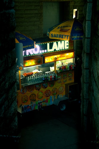 A brightly lit food cart featuring signs with images of various foods and drinks. Several umbrellas with branded logos provide shade while an array of bottled beverages is neatly arranged on the counter. The cart is nestled between tall, dark stone walls, providing a cozy nook within an urban environment.