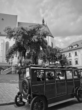 A vintage-style open bus with passengers is parked on a cobblestone street beside a church. The background features historic architecture with a prominent stone facade and a large tree providing shade. The scene has a timeless, nostalgic feel.