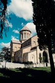 A historic stone church with a domed roof stands under a bright blue sky filled with fluffy white clouds. The architecture features elegant arches and intricate stonework. Tall trees and neatly trimmed grass surround the church, creating a peaceful atmosphere.