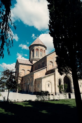 A historic stone church with a domed roof stands under a bright blue sky filled with fluffy white clouds. The architecture features elegant arches and intricate stonework. Tall trees and neatly trimmed grass surround the church, creating a peaceful atmosphere.