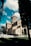 A historic stone church with a domed roof stands under a bright blue sky filled with fluffy white clouds. The architecture features elegant arches and intricate stonework. Tall trees and neatly trimmed grass surround the church, creating a peaceful atmosphere.