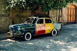 A vintage car painted with large blocks of green, red, and yellow sits parked on a cobblestone street. The word 'SIGNAGHI' is painted in white letters on the car's side. A large clay pot and tree branches rest on the car's roof, partially covered by the overhanging branches of a tree above. The surrounding area includes a stone and brick wall and a wooden gate.