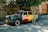 A vintage car painted with large blocks of green, red, and yellow sits parked on a cobblestone street. The word 'SIGNAGHI' is painted in white letters on the car's side. A large clay pot and tree branches rest on the car's roof, partially covered by the overhanging branches of a tree above. The surrounding area includes a stone and brick wall and a wooden gate.