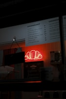 A dimly lit bakery or cafe interior features a prominent neon sign in the shape of a croissant glowing in red. A menu board lists various croissant options with prices. The surroundings include shelves with boxes and cups, suggesting a small and cozy setup.
