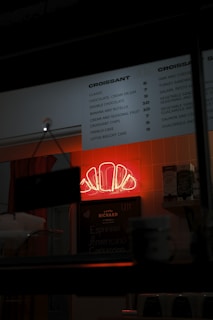 A dimly lit bakery or cafe interior features a prominent neon sign in the shape of a croissant glowing in red. A menu board lists various croissant options with prices. The surroundings include shelves with boxes and cups, suggesting a small and cozy setup.