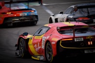 Three race cars are speeding down a track in close competition. A prominent red and yellow sports car is at the forefront with detailed branding and sponsorship decals visible. Behind it, two other cars are engaged in the race, each with distinctive racing liveries and designs. The image captures the dynamic and intense atmosphere of a professional car race.