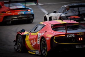 Three race cars are speeding down a track in close competition. A prominent red and yellow sports car is at the forefront with detailed branding and sponsorship decals visible. Behind it, two other cars are engaged in the race, each with distinctive racing liveries and designs. The image captures the dynamic and intense atmosphere of a professional car race.