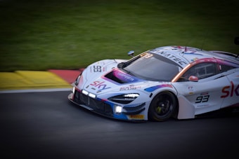 A sleek, high-performance race car is speeding along a track. The vehicle is adorned with various sponsor logos and features a number 93 on its side. The design includes vibrant accents of red, blue, and purple on its predominantly white body. The background shows a grassy area next to the asphalt track, adding to the dynamic racing atmosphere.