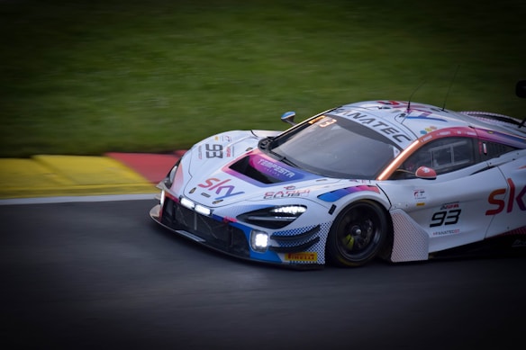 A sleek, high-performance race car is speeding along a track. The vehicle is adorned with various sponsor logos and features a number 93 on its side. The design includes vibrant accents of red, blue, and purple on its predominantly white body. The background shows a grassy area next to the asphalt track, adding to the dynamic racing atmosphere.